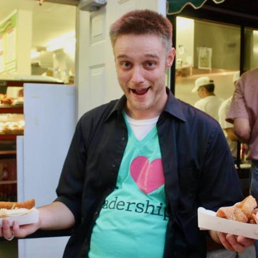 A younger Brian makes an excited face while holding food items and wearing a green shirt under a black button-up shirt. The green shirt says "I heart leadership".