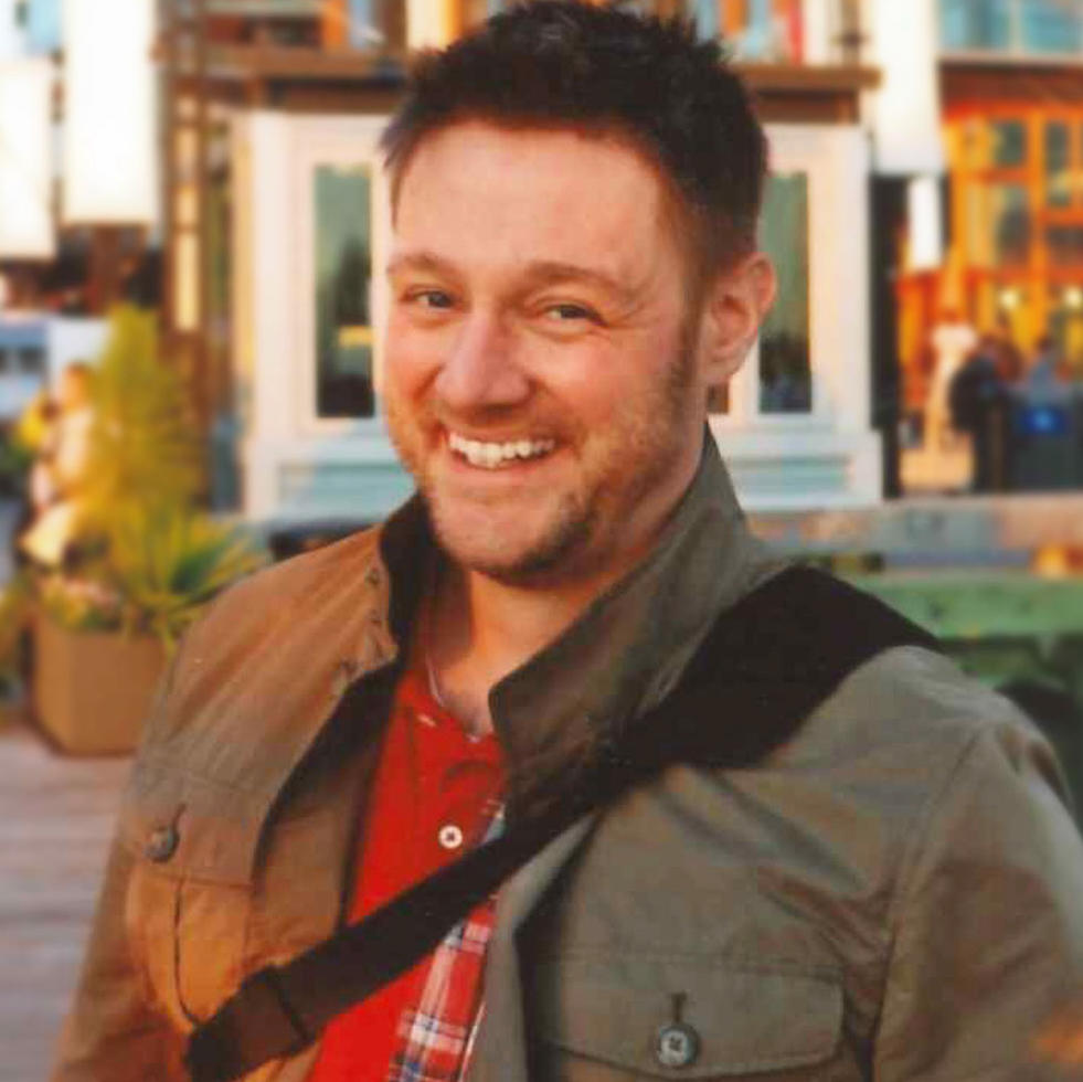 A close-up photo of Brian smiling while standing outside in a town square during a warm sunset. Brian wears a red shirt, brown jacket and cross-body bag.