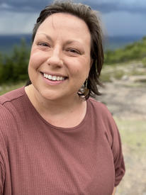 Laura, a white woman with brown hair, is smiling at the camera with her head tilted to the side.  Blurred behind her gray stone ground, green trees and a dark blue sky.
