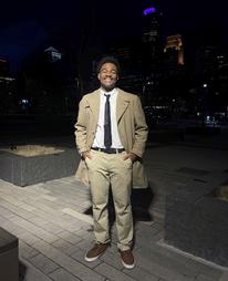 Julius wears a tan suit and smiles while standing in front of the city at night
