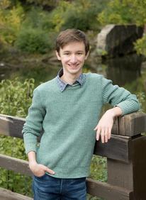 Close up photo of Aidan smiling while wearing a green sweater and resting his arm on a wooden bridge in front of a river