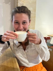 Close up photo of Ellery wearing a white shirt and holding a white coffee mug close to her face while smiling