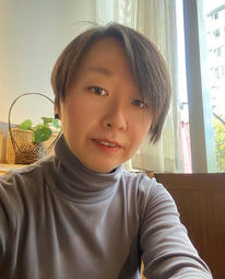 Close up photo of Yilin wearing a grey turtleneck sitting next to a window and in front of a houseplant