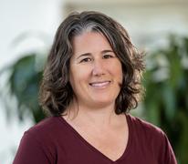 Close up photo of Jennifer Trost, a white woman with brown hair and a grey streak, wearing a maroon shirt with green plants in the distance background