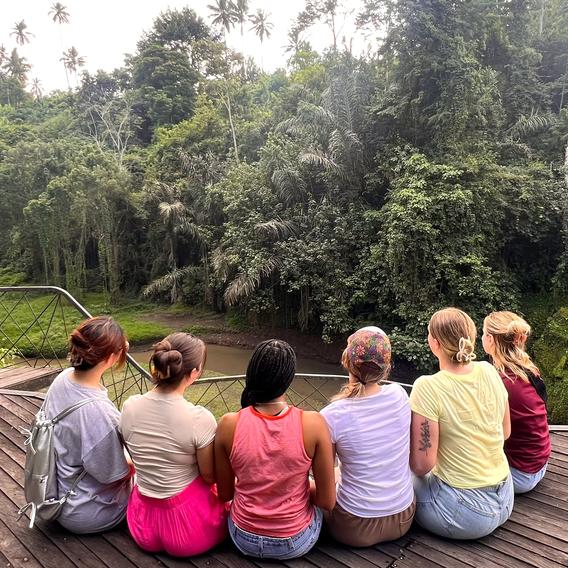 Close up photo from behind six students sitting in a semicircle on a wooden platform overlooking a lush jungle in Indonesia 