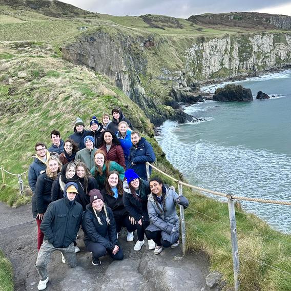 20 students wearing coats and hats stand in a group and smile along the cliffside. The cliff is covered in green grass and the water below is light blue.